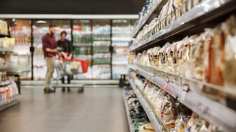 Rechts im Fokus ist ein Regal im Gang eines Supermarkts, das mit verschiedenen Produkten befüllt ist. Im Hintergrund stehen zwei Personen neben einem teilweise gefüllten Einkaufswagen. Hinter ihnen ist ein Kühlregal