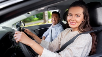 Am Steuer eines Wagens sitzt eine lächelnde Person mit offenen, dunklen Haaren, die direkt in die Kamera blickt. Die Person hat beide Hände am Lenkrad. Neben ihr sitzt eine Person, die zu ihr blickt und lächelt