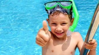 Ein Kind mit Taucherbrille und Schnorchel hält sich an einer Metallleiter am Rand eines Schwimmbeckens fest. Das Kind streckt die rechte Hand mit Daumen nach oben nach vorne. 