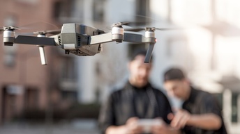 Im Vordergrund ist in der Luft eine schwebende Drohne. Im Hintergrund verschwommen stehen zwei Personen nebeneinander. Die linke hält die Fernsteuerung der Drohne.