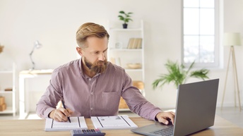 Frontale Aufnahme einer Person, die an einem Tisch sitzt und auf einen aufgeklappten Laptop blickt. Vor der Person sind nebeneinander zwei Stapel Papiere. Daneben steht ein Taschenrechner