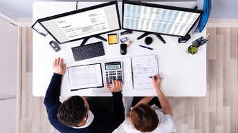 Top-Shot zweier nebeneinander an einem Schreibtisch sitzender Personen. Auf dem Tisch sind zwei Bildschirme. Auf dem linken steht Invoice. Vor den Bildschirmen sind Unterlagen, auf die die beiden Personen blicken