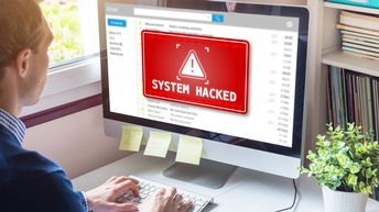 Person sitzt vor einem Computer bei einem Schreibtisch und die Meldung System Hacked erscheint am Display in roter Warnung