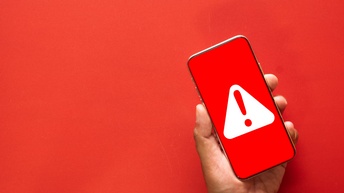 Die Hand einer Person hält ein Handy, dessen Hintergrund rot ist. Auf dem Bildschirm ist ein rotes Rufzeichen in einem weißen Dreieck