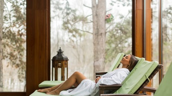 Person mit weißem Bademantel liegt auf einer Liege mit grüner Auflage in einem Wellnessbereich mit gläsernen Fronten und Blick in die Natur