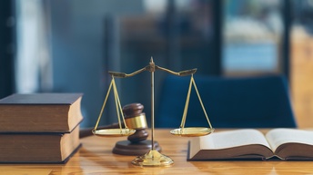 Bücher sowie ein Richterhammer, eine goldene Waage und ein aufgeschlagenes Buch auf einem Holztisch