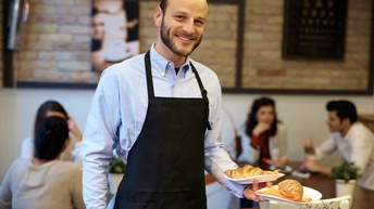 Lächelnde Person in schwarzer Schürze hält in einer Hand zwei Teller mit Croissants, im Hintergrund verschwommen weitere Personen an Tischen sitzend