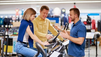 Mitarbeiter eines Sportartikelhandels berät Pärchen bei Kauf eines Fitnessgeräts