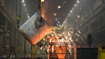 In einer großen Halle hängt von der Decke ein sehr großer Stahlbehälter, aus dem eine rötliche Flüssigkeit in einen anderen stehenden Behälter fließt. Neben den beiden Behältern steht eine Person mit einem Gesichtsvisier und dicken Handschuhen