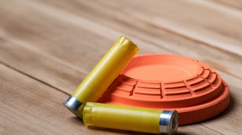 Orange Wurfscheibe aus Ton auf Holzuntergrund liegend, darauf und daneben zwei gelbe Patronen