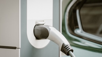 Nahaufnahme einer E-Ladestation, in der ein Stecker steckt. Im Hintergrund parkt ein Auto
