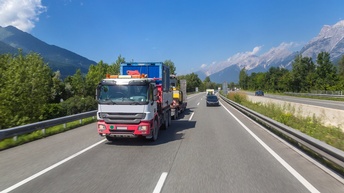 LKW und Autos fahren auf einer Autobahn, im Hintergrund sieht man abseits einen blauen Himmel sowie Bäume und Berge 