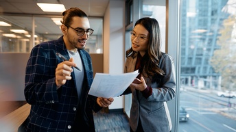 Zwei Personen stehen nebeneinander im Gang eines Büros. Die linke Person hält Zettel, auf die beide blicken