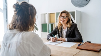 Zwei Personen sitzen sich gegenüber an Tisch in Büro, im Hintergrund Regal und Uhr