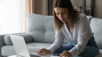 Person in gestreiftem Hemd arbeitet mit einem Laptop und rechnet mit einem Taschenrechner während sie auf einem Sofa sitzt