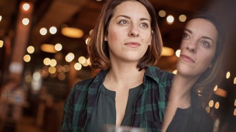 Portrait von einer Person mit schulterlangen braunen Haaren, grüner Bluse und grünem kariertem Hemd vor einem beleuchteten Restaurantambiente.