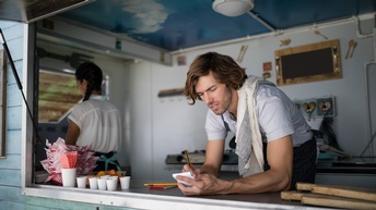 Person mit längeren braunen Haaren und Bart hat ein Geschirrtuch übergeworfen, steht in einem Foodtruck und notiert sich mit einem Stift etwas auf einem Notizblock, eine weitere Person steht ebenfalls im Anhänger