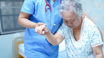 Person in blauem Arbeitsgewand mit rotem Stethoskop um den Hals stützt ältere Person in Nachthemd