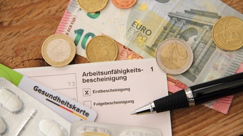 Top-Shot von Münzen, Geldscheinen, einem Zettel, auf dem Arbeitsunfähigkeitsbescheinigung steht, einer Plastikkarte, auf der Gesundheitskarte steht, und Tabletten auf einer hölzernen Unterfläche.