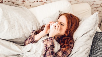 Eine Person liegt auf einer hellen Unterfläche. Die Person trägt ein kariertes Hemd und hat rote Haare. Über ihr ist eine hellgraue Decke. Sie hält sich mit beiden Händen ein Taschentuch vor die Nase. Ihre Augen sind geschlossen