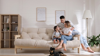 Auf einem hellen Sofa sitzen ein Kind und zwei erwachsene Person. Vor dem Sofa liegt ein Hund. Das Kind hält ein aufgeklapptes Buch. Die erwachsenen Personen schauen auf das Kind. Das Kind schaut auf die erwachsenen Personen