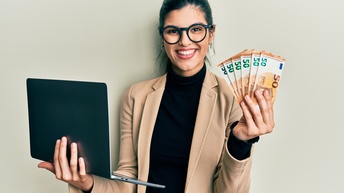 Halbporträt einer lächelnden Person in einem schwarzen Oberteil mit Sakko und Brillen, die vor einer hellen Wand steht. In der rechten Hand hält sie einen aufgeklappten Laptop. In der linken Hand hält sie mehrere Fünfzig-Eurogeldscheine.
