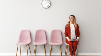 In einem Raum stehen vier Stühle. Auf dem Stuhl ganz rechts sitzt eine Person in einem roten Anzug. Sie hält Unterlagen in den Händen. An der Wand ist eine Wanduhr montiert