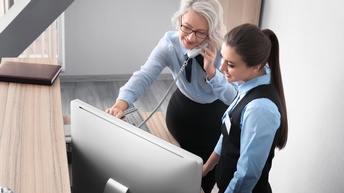 Zwei Personen in Uniform stehen hinter einem Tresen aus hellem Holz. Beide tragen Uniform. Die rechte hat an der Weste einen Ausweis. Die linke telefoniert. Beide blicken auf den Bildschirm