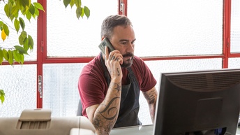 Eine Person sitzt an einem Tisch und telefoniert. Vor ihr ist ein Computer. Neben dem Computer ist ein Drucker