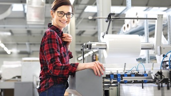 Eine Person mit Brille steht seitlich vor einer großen Maschine. Auf einer Fläche der Maschine liegt ein hoher, weißer Papierstapel. Schräg über dem Stapel ist eine Metallrolle, auf der eine dicke, weiße Rolle ist. Die Person blickt direkt in die Kamera.