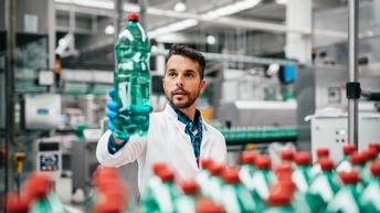 Person mit Gummihandschuhen in weißem Kittel steht in Werkshalle mit Fließbändern und hält grüne Plastikflasche mit Flüssigkeit gefüllt und blickt darauf, im Vordergrund verschwommen zahlreiche Flaschen