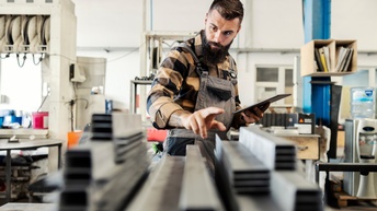 Person in kariertem Hemd und Arbeitskleidung steht in einer Werkstatt und kontrolliert Betriebsmittel während ein Tablet in der Hand gehalten wird