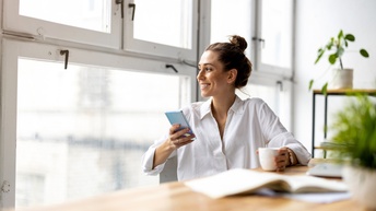 Lächelnde Person blickt aus Fenster an Tisch sitzend mit Smartphone in der Hand