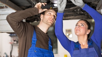 Zwei Personen in blauen Latzhosen stehen nebeneinander unter einem Fahrzeug, das auf einer Hebebühne ist. Die rechte Person trägt Handschuhe und hantiert am Unterboden des Fahrzeugs. Die linke Person hält eine Taschenlampe an diese Stelle