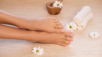 Nahaufnahme der liegenden Füße einer Person. Um die Füße liegen weiße Blüten, drei eingerollte Handtücher und ein hölzerner Mörser. Die Zehen der Person haben einen weißen Rand. Auf den Zehen ist durchsichtiger Nagellack.