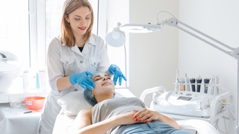 Eine Person liegt auf einer Liege und trägt ein Haarnetz. Am Kopfende steht eine Person in einem weißem Kittel. Sie trägt mit blauen Handschuhen eine weiße Substanz auf dem Gesicht der liegenden Person auf