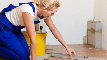 Seitliche Aufnahme einer Person in blauer Latzhose, die auf einem hellbraunen Boden kniet. Links neben ihr sind Fliesen, auf die sie eine Wasserwaage hält, auf die sie blickt. Links daneben steht ein gelber Kübel.