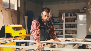 Eine Person mit Schutzbrille steht in einer Werkstatt zwischen einem großen Rahmen aus Holzbalken, auf den sie den Plastikrahmen hält.