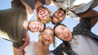 Aufnahme von unten von fünf Personen, die einen Kreis bilden und ihre Köpfe zusammenstecken. Die Köpfe der Personen treffen in der Mitte aufeinander. Die Personen blicken nach unten zur Kamera