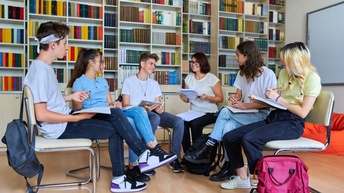 Eine Gruppe von Schüler:innen sitzt in einem Halbkreis in einer Bibliothek mit einer Lehrperson und spricht miteinander.