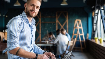 Person in blau-weiß gestreiften Hemd lächelt und hat die Hände auf der Laptoptastatur, im Hintergrund weitere Personen am Tisch sitzend