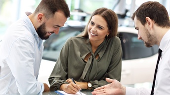 Drei lächelnde Personen, eine Hält Stift auf Dokument, im Hintergrund Auto