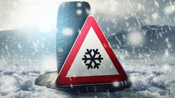 Ein Autoreifen ist  auf einer schneebedeckten Straße aufgestellt. Vor dem Reifen ist ein dreieckiges Schild, auf dem eine Schneeflocke ist. Es herrscht Schneeregen, im Hintergrund ist eine Berglandschaft zu erkennen. 