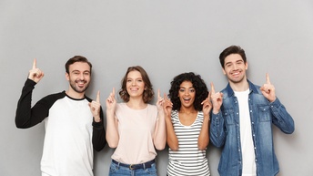 Vier Personen in Jeans und T-Shirts stehen nebeneinander vor einer grauen Wand und lächeln. Jede Person blickt in die Kamera und deutet mit beiden Zeigefingern jeweils in die Luft