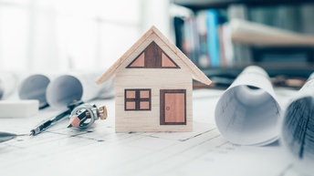 Ein kleines Modellhaus aus Holz steht auf einem bedruckten Papier. Rechts und links neben dem Haus sind mehrere Papierrollen. Links neben dem Haus sind ein Stift und ein Zirkel.