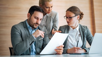 Zwei Personen in Bürokleidung sitzen nebeneinander an einem Tisch. Hinter ihnen steht eine weitere Person. Die links sitzende Person hält einen Zettel. Alle drei blicken auf den Zettel