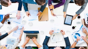 Personen in Businesskleidung sitzen rund um einen Besprechungstisch mit Laptops, statistischen Unterlagen, Notizblöcken, Tablets, Tassen Kaffee, Gläsern Wasser sowie Stiften, Topview