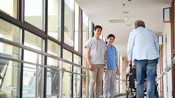 In einem Gang stehen in der linken Hälfte zwei Personen in Kitteln. Diese sind zur Kamera gewandt. In der rechten Hälfte ist die Rückenansicht einer anderen Person, die einen Rollstuhl schiebt