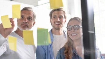 Drei Personen vor transparenter Tafel versammelt, auf der Post its kleben