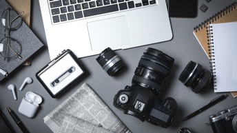 Topshot eines grauen Tisches, auf zwei Kameras, verschiedene Objektive, Kopfhörer, ein Laptop, zwei Zeitungen, zwei Notizbücher, eine Brille, ein Smartphone, ein Notizbuch und eine Brille liegen